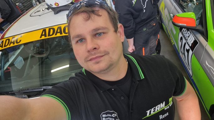 Selfie-Mode Mann mit einem Polo-Shirt steht neben einem Rennwagen in einer Werkstatt.