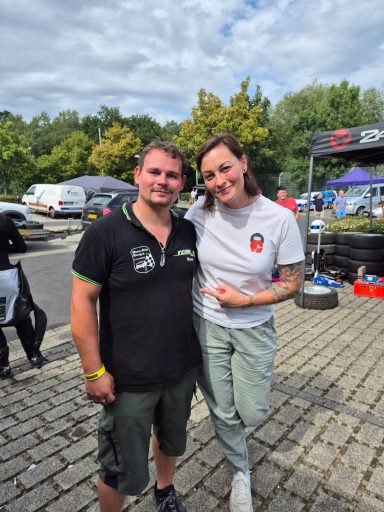 René mit Lina van de Mars Zwei Personen lächeln, stehen im Freien vor Autos und Reifen, umgeben von Bäumen.