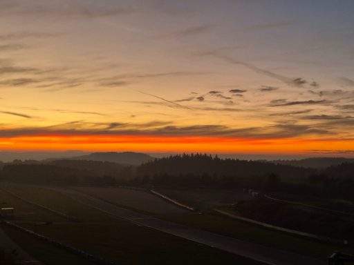 Sonnenaufgang vor dem 3h-Rennen Sonnenuntergang über einer hügeligen Landschaft mit orangefarbenem Himmel.