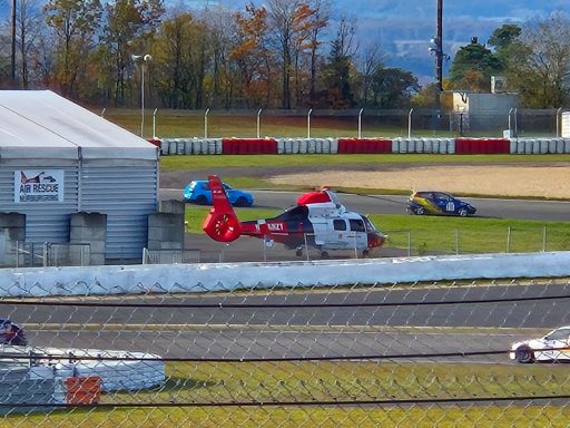 Rettungshelikopter Roter Hubschrauber neben Rennautos auf einer Rennstrecke, im Hintergrund Bäume im Herbst.