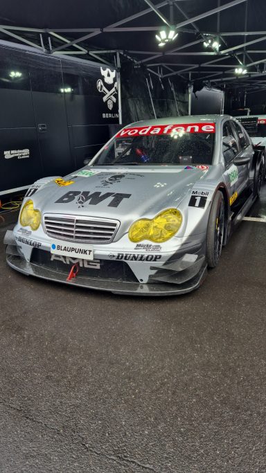 Der DTM Legends Mercedes Silberner Rennwagen mit gelben Scheinwerfern und Sponsorenaufklebern in einer Box.