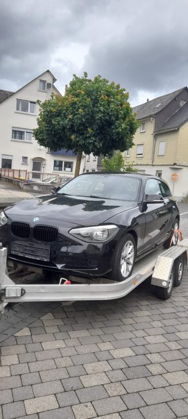Neuestes Teammitglied Schwarzes Auto auf einem Anhänger, umgeben von Wohnhäusern und einem Baum.