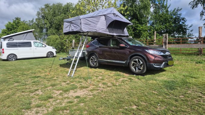 Renés Schlafplatz Ein SUV mit einem Dachzelt auf einem grünen Rasenplatz, umgeben von Bäumen.